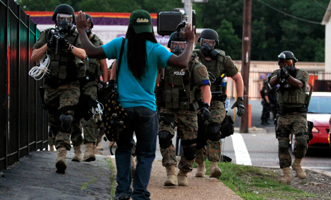 police dressed as soldiers, with automatic rifles, body armor and gas masks, aim their weapons at a black man